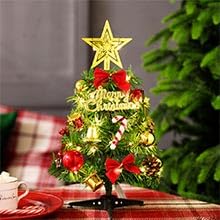 Small tabletop Christmas tree with gold star topper, ornaments, and lights. Set on wooden surface with gift boxes in background.